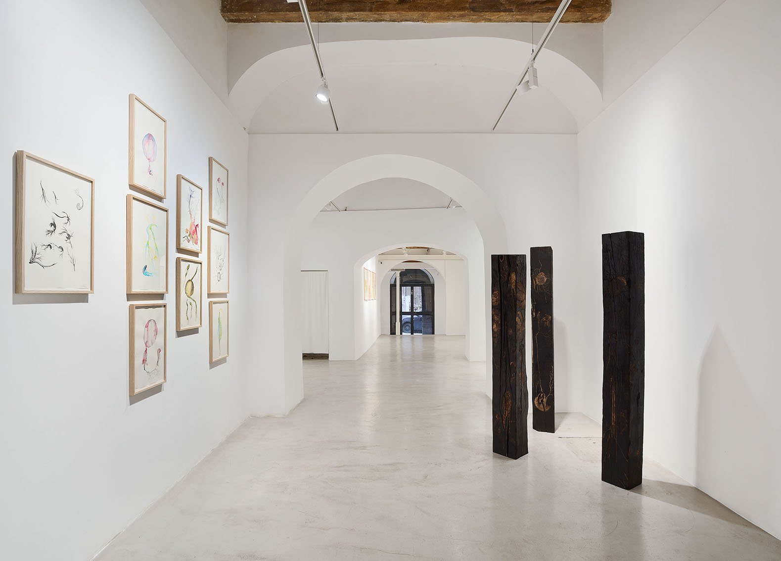 White-walled art gallery with framed drawings lining the left wall, dark vertical sculptures on the right, and arched passageways in the distance.