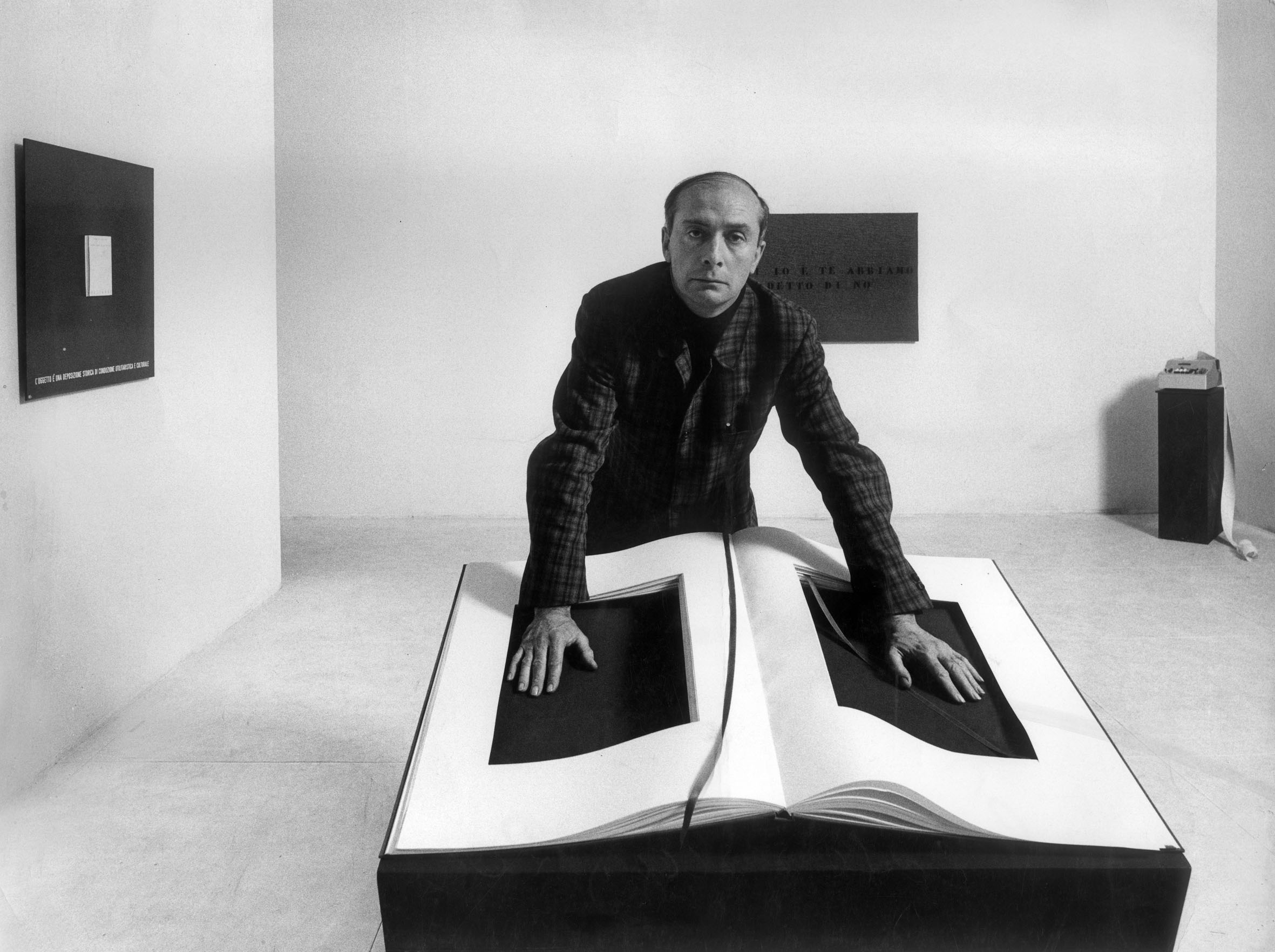 Man in plaid shirt leaning over oversized open book sculptures in a gallery.