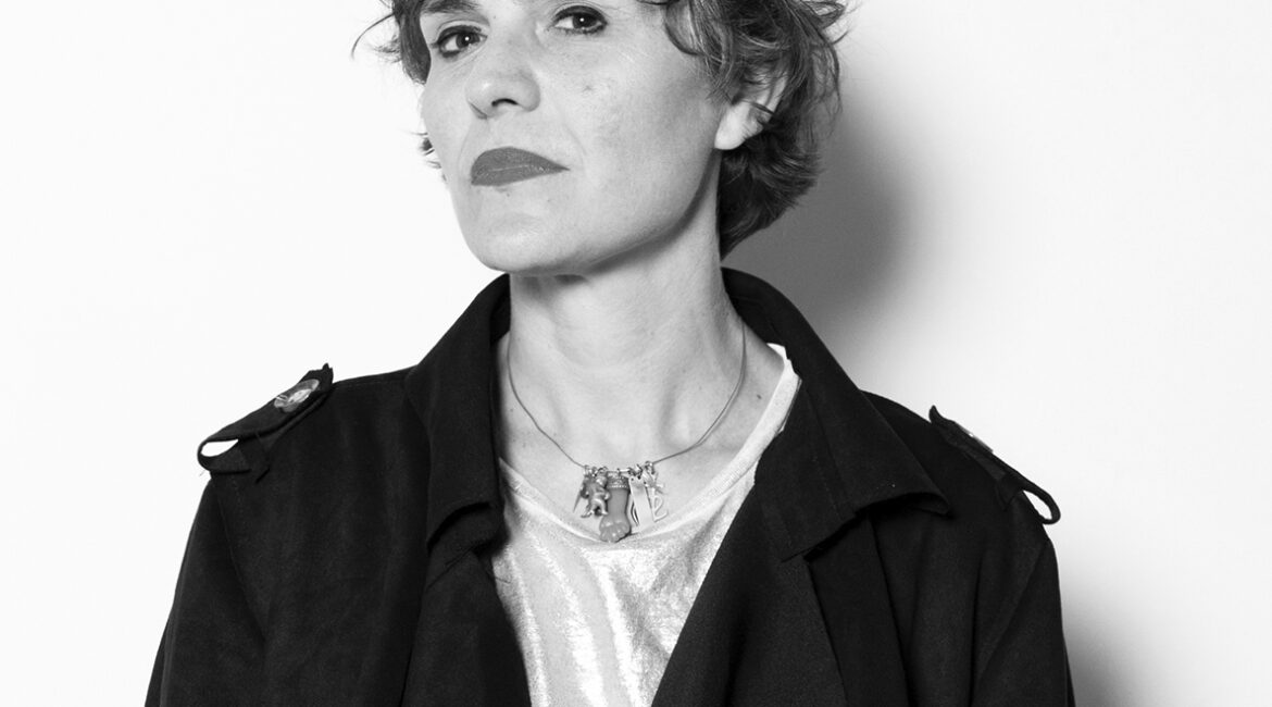 Black-and-white head-and-shoulders portrait of a woman with short hair, wearing a dark coat and a necklace, looking at the camera with a confident expression.