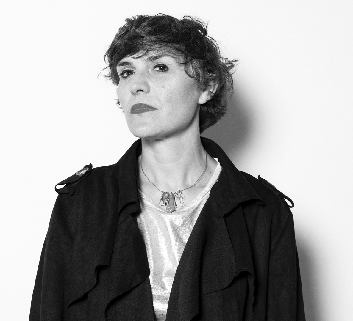 Black-and-white head-and-shoulders portrait of a woman with short hair, wearing a dark coat and a necklace, looking at the camera with a confident expression.