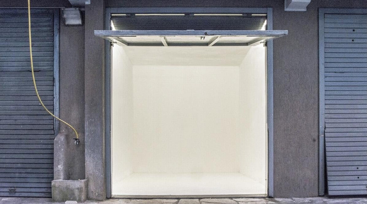 Open, empty white box-like doorway set in a gray wall, with blue-gray shutters on both sides and a yellow hose along the left column outside.