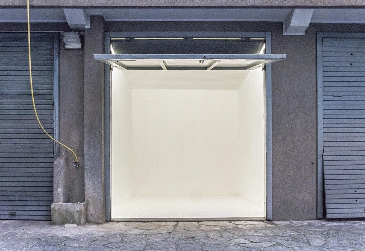 Open, empty white box-like doorway set in a gray wall, with blue-gray shutters on both sides and a yellow hose along the left column outside.