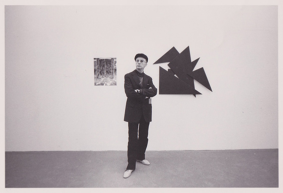 Man in a suit and flat cap stands with arms crossed in a white-walled gallery, next to a small framed artwork and a large black geometric wall sculpture.