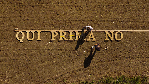 Aerial view of two people standing on a dirt field beside large wooden letters laid out on the ground, spelling a phrase.